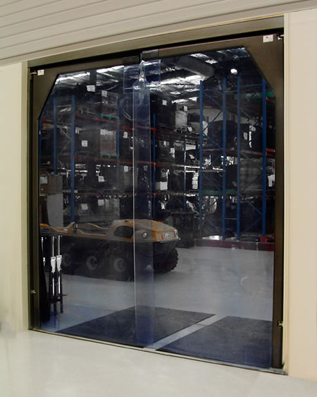 Transparent industrial strip curtain door leading to a warehouse with shelves and a yellow vehicle in the background.