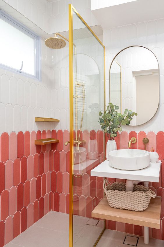 Modern bathroom with pink and white tiled walls, gold fixtures, glass shower, round mirror, vessel sink, and potted plant.