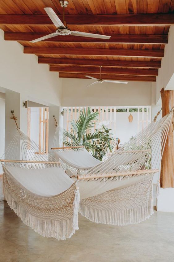 Cozy indoor space with multiple white hammocks, wooden ceiling, fans, and lush green plants, perfect for relaxation.