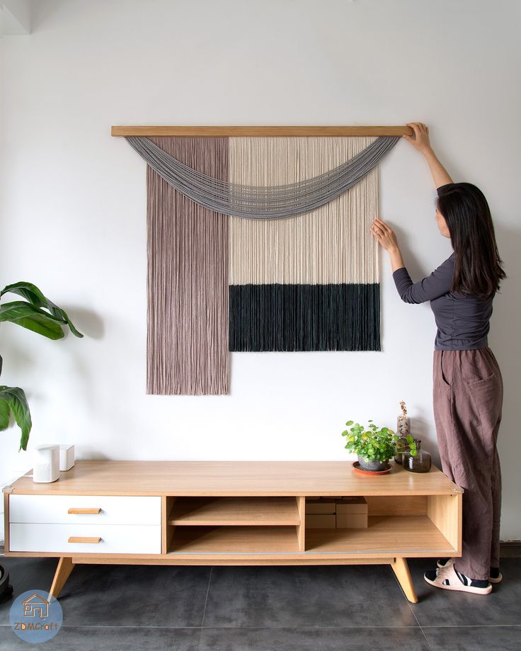 Woman hanging a modern macramé wall art above a minimalist wooden TV stand with plants, adding chic decor to the living room.