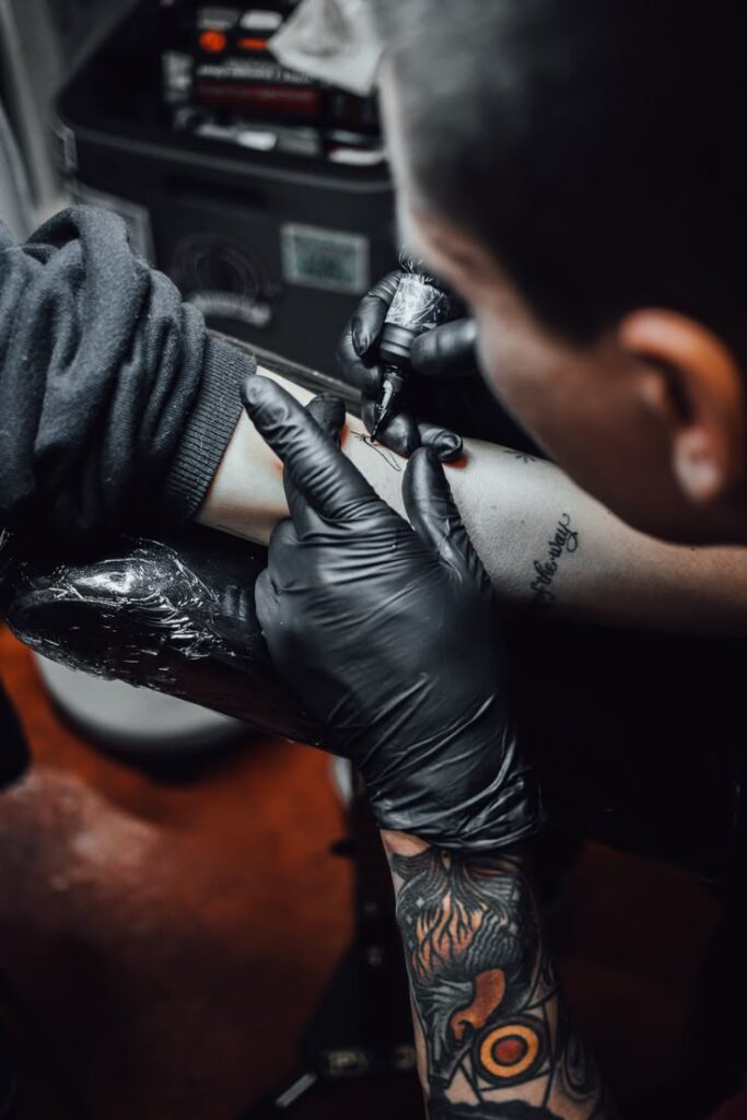 LOST CAUSE FLASH TATTOO TOUR Tattoo artist wearing black gloves working on a forearm tattoo design in studio, showcasing intricate skill and precision. | Sky Rye Design Tattoo artist wearing black gloves working on a forearm tattoo design in studio, showcasing intricate skill and precision.