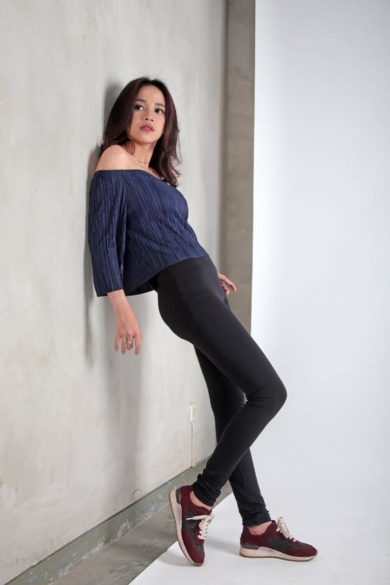 Woman modeling casual outfit with purple off-the-shoulder top, black leggings, and maroon sneakers, leaning on a concrete wall.