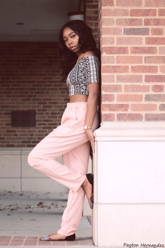 Woman in patterned crop top and pink pants leans against a brick wall, showcasing chic, casual fashion.