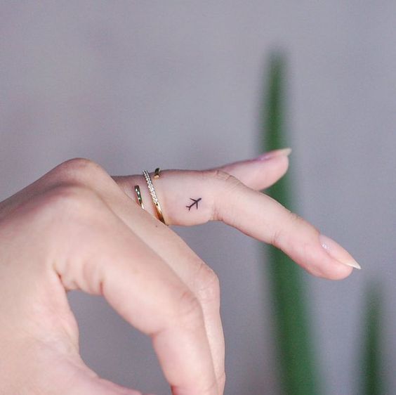 Fingertip airplane tattoo with gold rings on woman's hand, minimalist travel design.