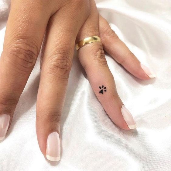 Close-up of a woman's hand with a wedding ring and a small paw print tattoo on the pinky finger, polished nails.