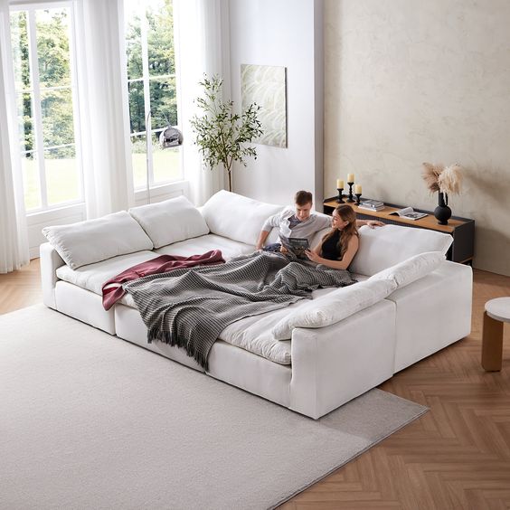 Cozy modern living room with a white sectional sofa, blanket, and two people reading by large windows with natural light.