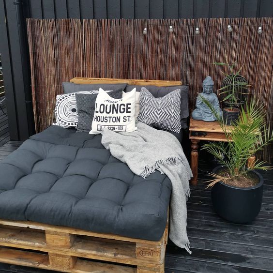 Outdoor lounge area with cozy pallet bed, gray cushions, decorative pillows, blanket, Buddha statue, and potted plant.