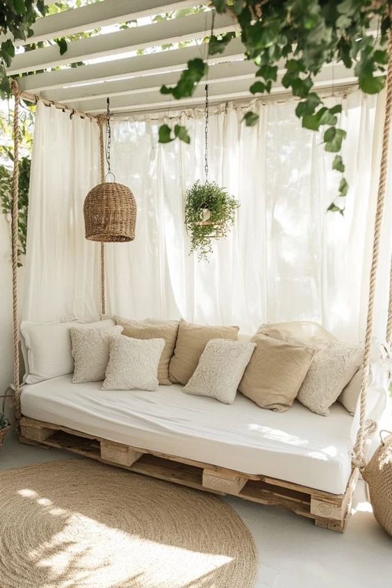 Cozy outdoor lounge area with pallet sofa, cushioned pillows, wicker lampshade, hanging plants, and white curtains.