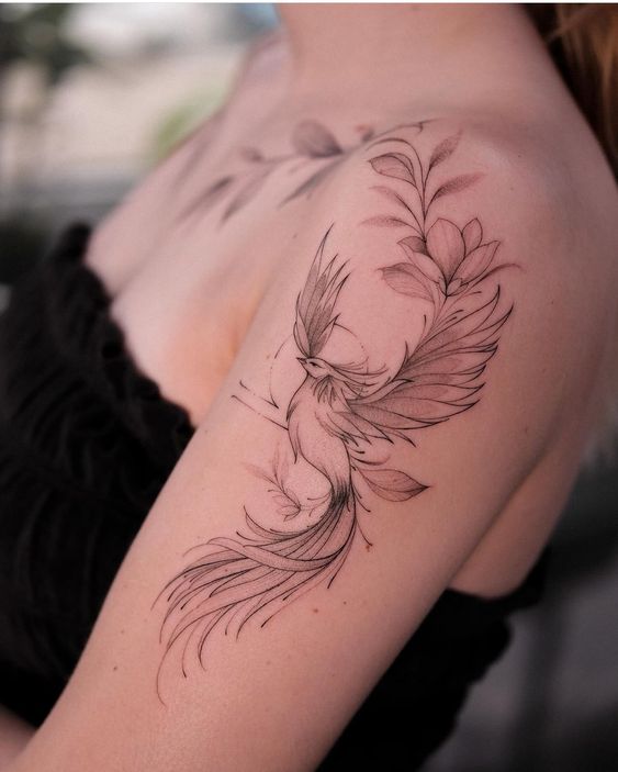 Delicate bird tattoo on woman's shoulder with detailed feathers and floral design, black ink.