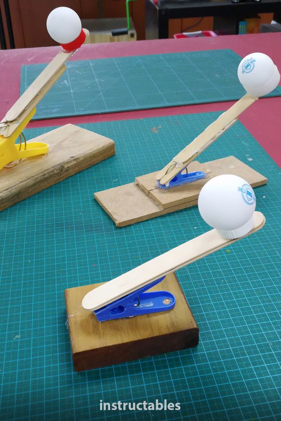 Three DIY catapults made from wooden bases, popsicle sticks, and clothespins, each launching a ping pong ball.