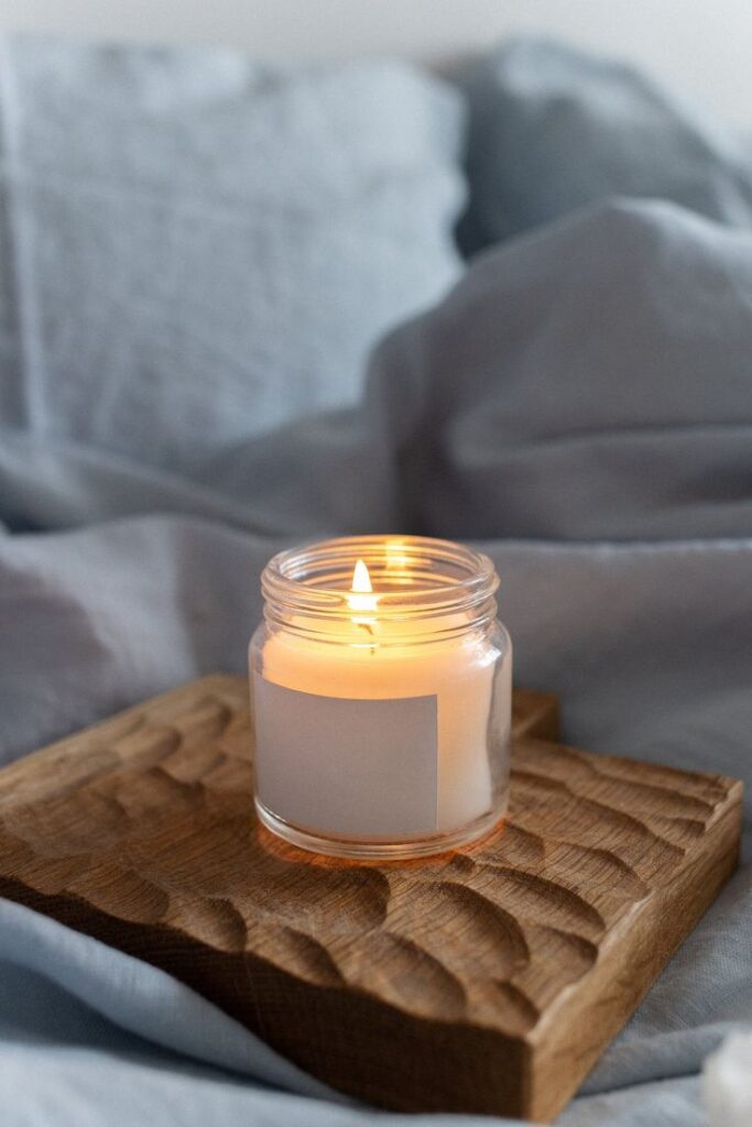 Lit scented candle in a glass jar on a wooden tray, creating a cozy and relaxing atmosphere with soft bedding in the background.