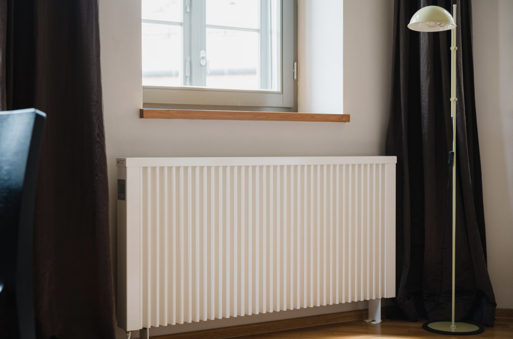 Modern white radiator underneath a window near curtains and a floor lamp, providing cozy home heating.