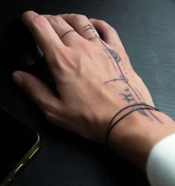 Hand with abstract linear tattoo using a computer mouse on a dark wooden desk, next to a smartphone.