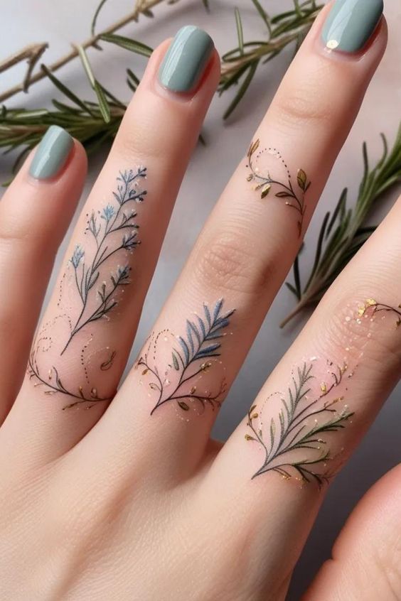 Close-up of fingers with intricate floral tattoos and sage green nail polish, complemented by fresh rosemary sprigs in the background.