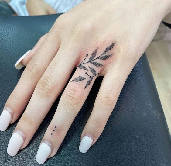 Hand with intricate leaf design tattoo on finger, next to simple black dot tattoo, and adorned with manicured nails.