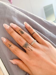 Close-up of a hand with rings and small intricate finger tattoos, resting on a grey fabric background.