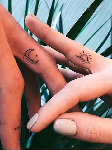 Close-up of two fingers with minimalistic moon and sun tattoos against a leafy background.