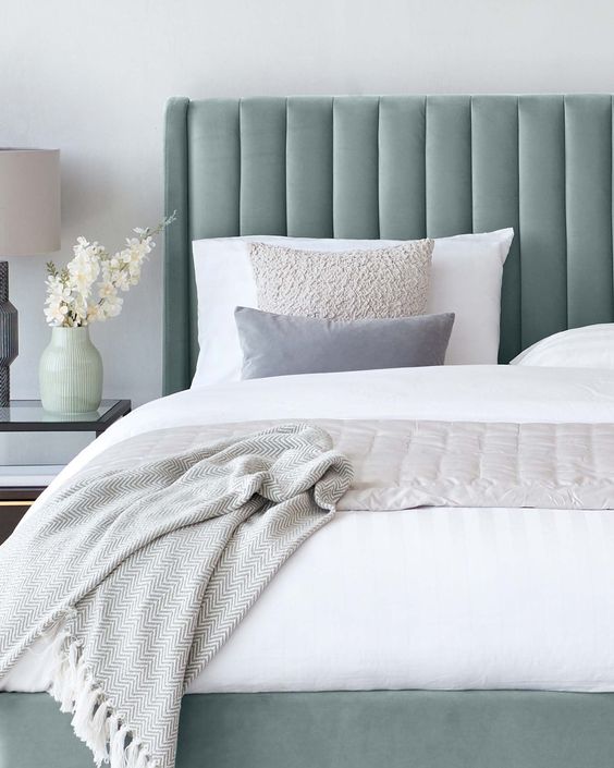 Elegant bedroom with gray headboard, textured throw pillows, and cozy blanket. Modern bedside table with lamp and vase of flowers.