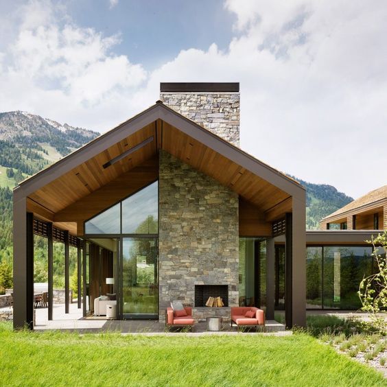 Modern house with a gable roof, stone fireplace, and large glass windows surrounded by mountains and green landscape.