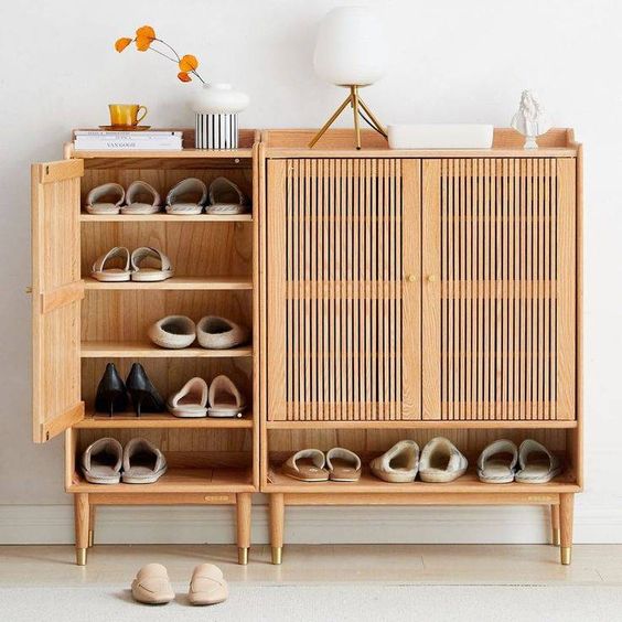 Modern wooden shoe cabinet with open shelves displaying various shoes and decorative items on top in a minimalist setting.