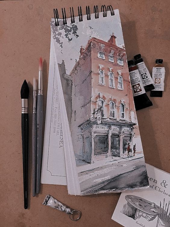 Watercolor painting of a building on a spiral sketchbook with brushes and paint tubes on a brown surface.