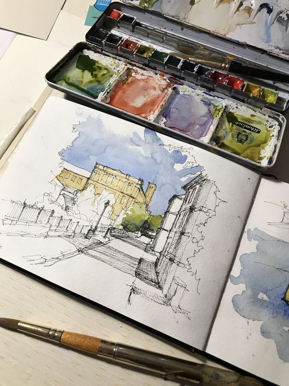 Watercolor sketch of a cityscape next to an open paint palette and brush on a white surface.