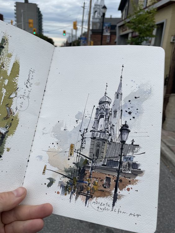 Hand holding a sketchbook with an artistic drawing of a church and street scene, cityscape in the background.