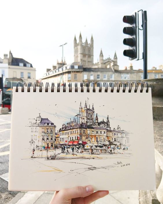 Hand holding a sketchbook with a drawing of a classic building matching the real scene in the background.