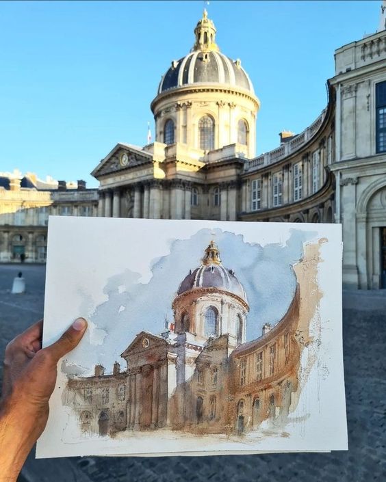 Hand holding a watercolor painting of a historic domed building, with the same building in the background under a blue sky.