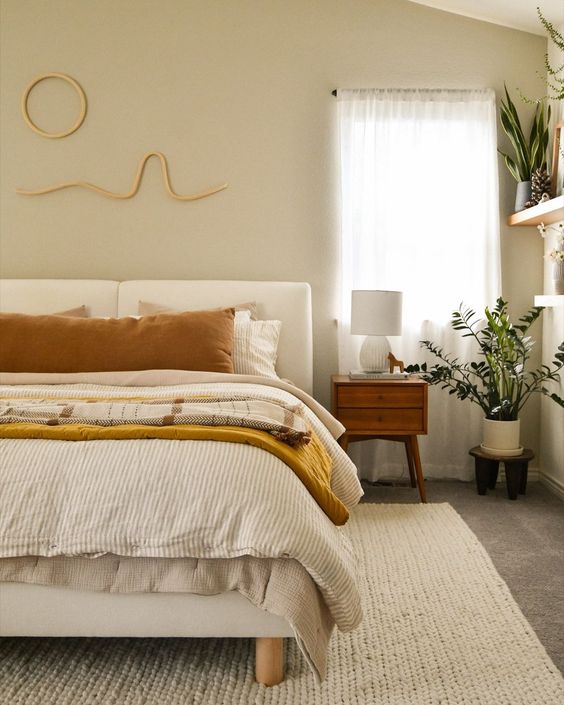 Cozy modern bedroom with neutral tones, natural light, and leafy plants next to a bedside table and lamp.