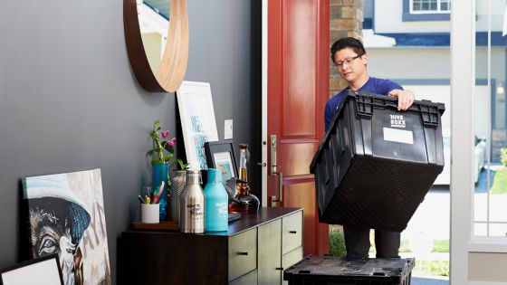 Man moving into a new home, carrying a black storage box through the front door, surrounded by household items and decor.