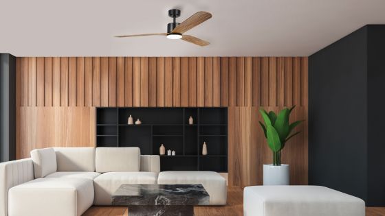 Modern living room with wood panel walls, white sectional sofa, black shelves, potted plant, ceiling fan, and black coffee table.