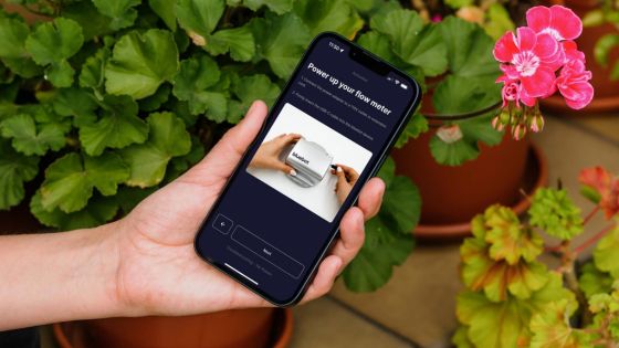 Hand holding a smartphone with a tech-related screen, surrounded by lush green and pink flowering plants.