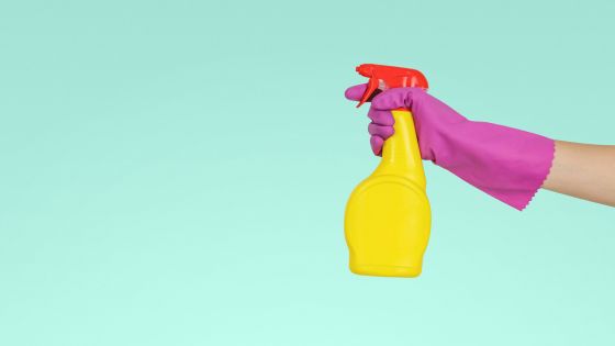 Person wearing pink gloves holding yellow spray bottle against light green background, symbolizing cleaning and sanitizing.