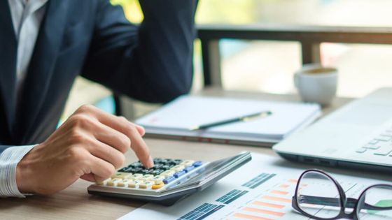 Business professional using a calculator for financial analysis at their desk with charts, laptop, and coffee in the background.