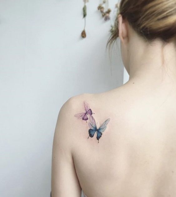 Woman with delicate butterfly tattoo on her upper back, featuring pink and blue butterflies, against a minimalistic background.