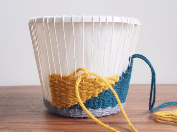 Partially completed yarn weaving project on a white paper cup, with yellow, blue, and grey yarn on a wooden surface.