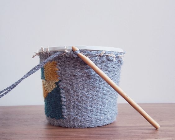 Gray crochet basket with wooden crochet hook on wooden table, featuring blue and yellow geometric shapes.