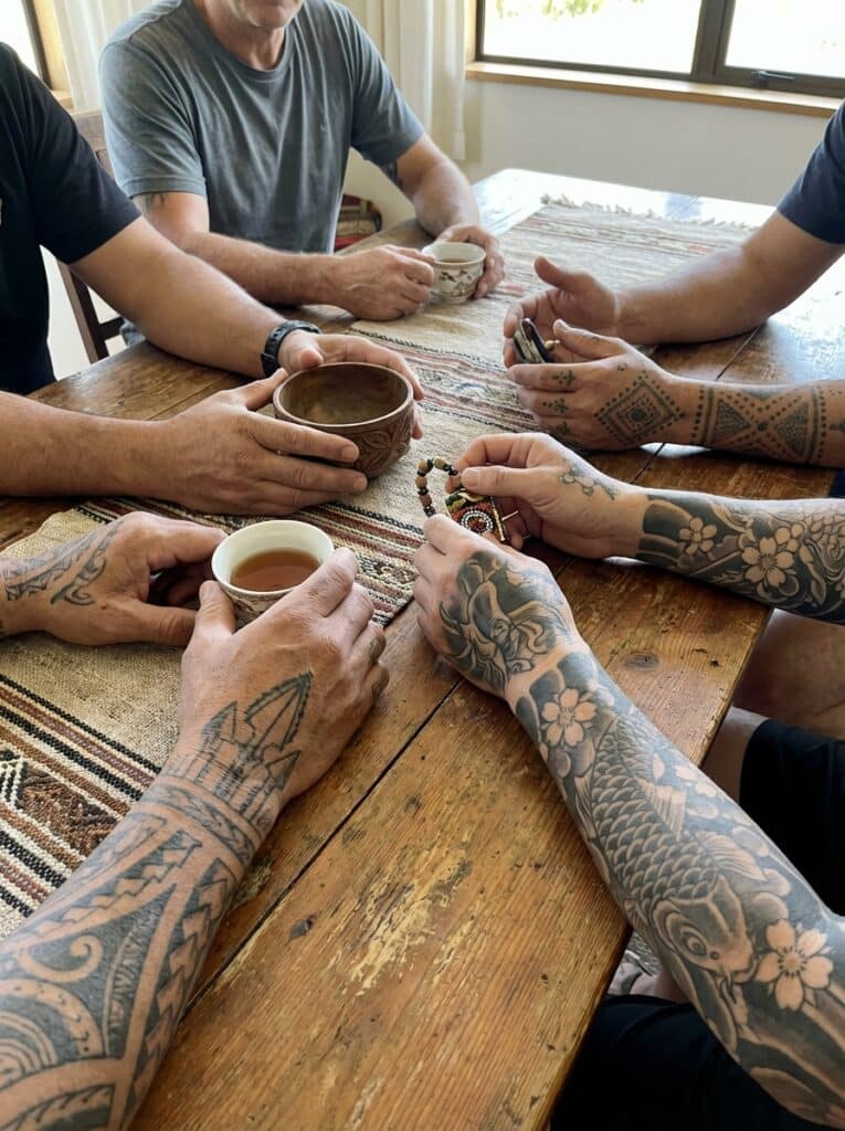 ai-photo-studio-1766759640084-2 Group of tattooed individuals sharing coffee and conversation around a rustic wooden table with a striped tablecloth. | Sky Rye Design Group of tattooed individuals sharing coffee and conversation around a rustic wooden table with a striped tablecloth.
