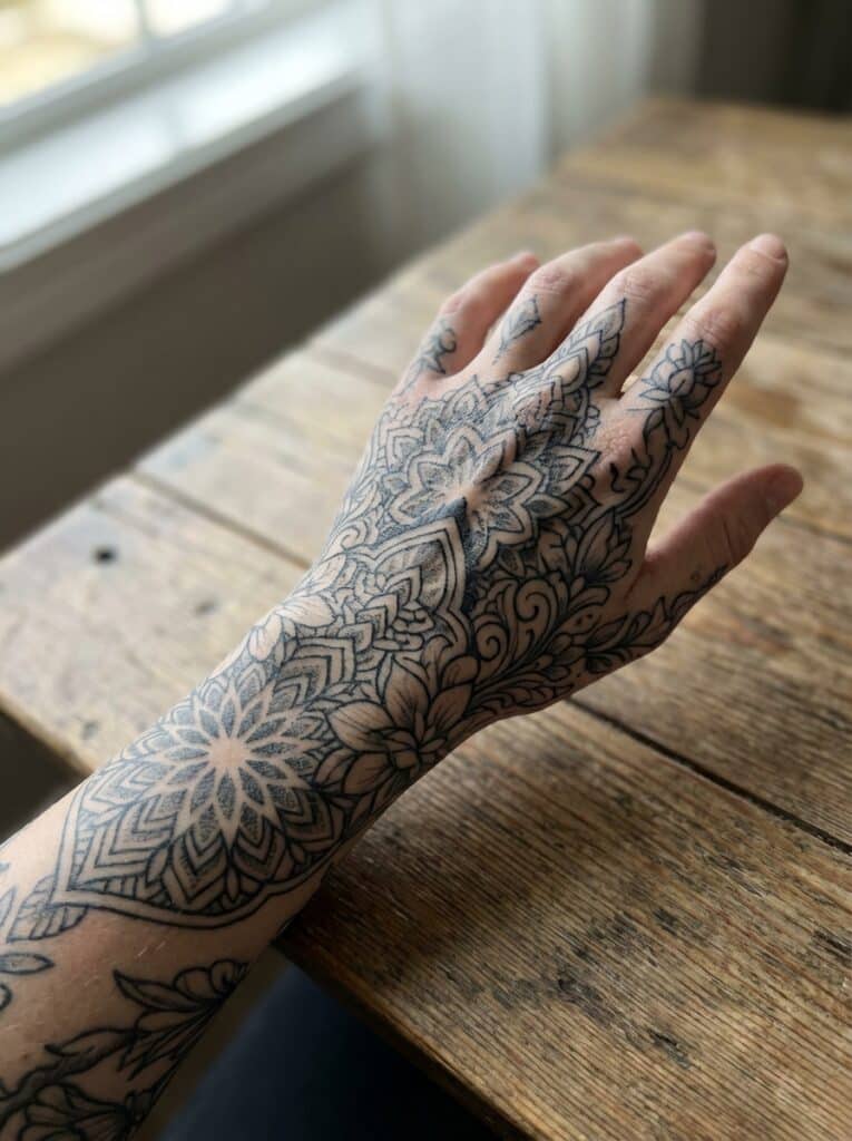ai-photo-studio-1766759767253-1 Intricate black floral mandala tattoo on hand and wrist, displayed on a wooden table, showcasing detailed artistry. | Sky Rye Design Intricate black floral mandala tattoo on hand and wrist, displayed on a wooden table, showcasing detailed artistry.