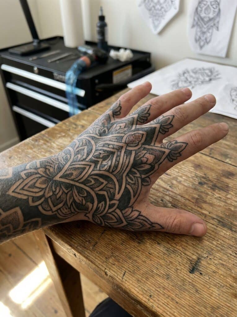 ai-photo-studio-1766759768853-2 Intricate floral tattoo design on a hand resting on a wooden table, showcasing detailed geometric patterns in a tattoo studio setting. | Sky Rye Design Intricate floral tattoo design on a hand resting on a wooden table, showcasing detailed geometric patterns in a tattoo studio setting.