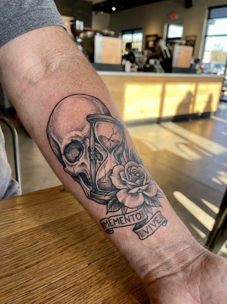 ai-photo-studio-1766760071986-2 Forearm tattoo featuring a skull, rose, hourglass, and Memento Vive text in a cozy cafe setting. | Sky Rye Design Forearm tattoo featuring a skull, rose, hourglass, and Memento Vive text in a cozy cafe setting.