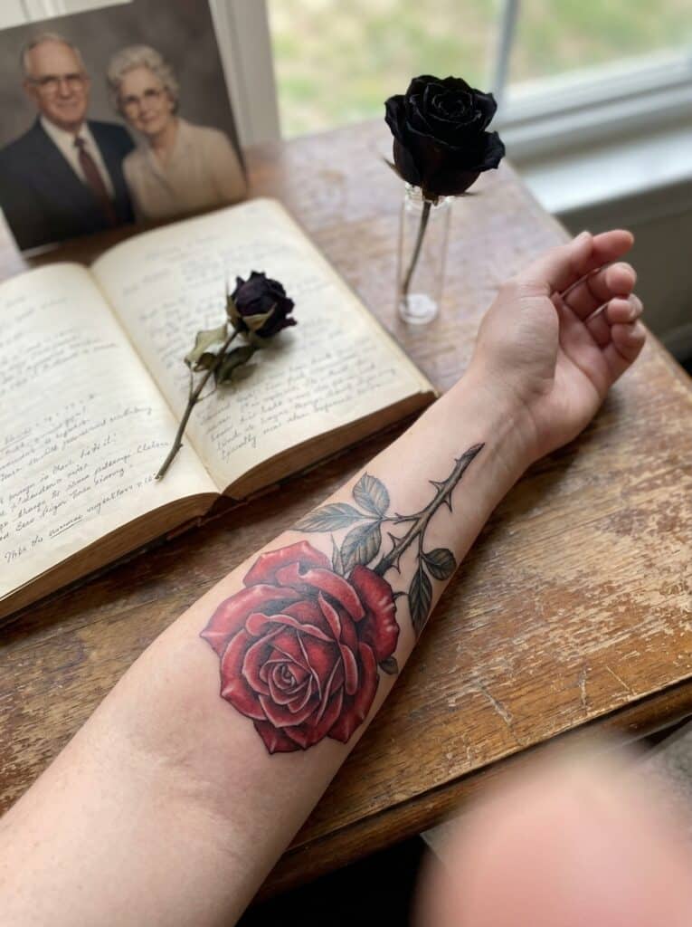 ai-photo-studio-1766760115635-1 Rose tattoo on forearm next to vintage photo, book, and dried roses on a wooden table by a window. | Sky Rye Design Rose tattoo on forearm next to vintage photo, book, and dried roses on a wooden table by a window.