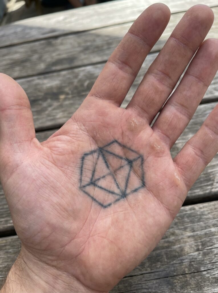ai-photo-studio-1766760175116-2 Geometric tattoo of a hexagon with lines on an open palm, set against a wooden table background. Minimalist design, hand tattoo art. | Sky Rye Design Geometric tattoo of a hexagon with lines on an open palm, set against a wooden table background. Minimalist design, hand tattoo art.