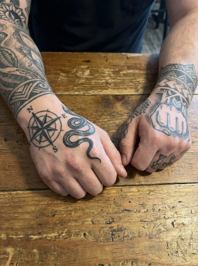 ai-photo-studio-1766760791053-1 Tattooed hands with compass and snake design on a wooden table, showcasing intricate ink art and geometric patterns. | Sky Rye Design Tattooed hands with compass and snake design on a wooden table, showcasing intricate ink art and geometric patterns.