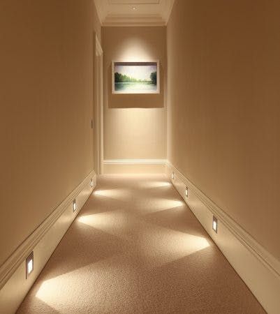 Dimly lit hallway with wall-mounted lights, beige carpet, and a landscape painting at the end.