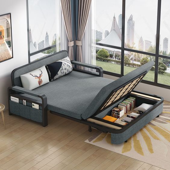 Modern grey convertible sofa bed with storage space, accented pillows, and a large window view of cityscape and greenery.