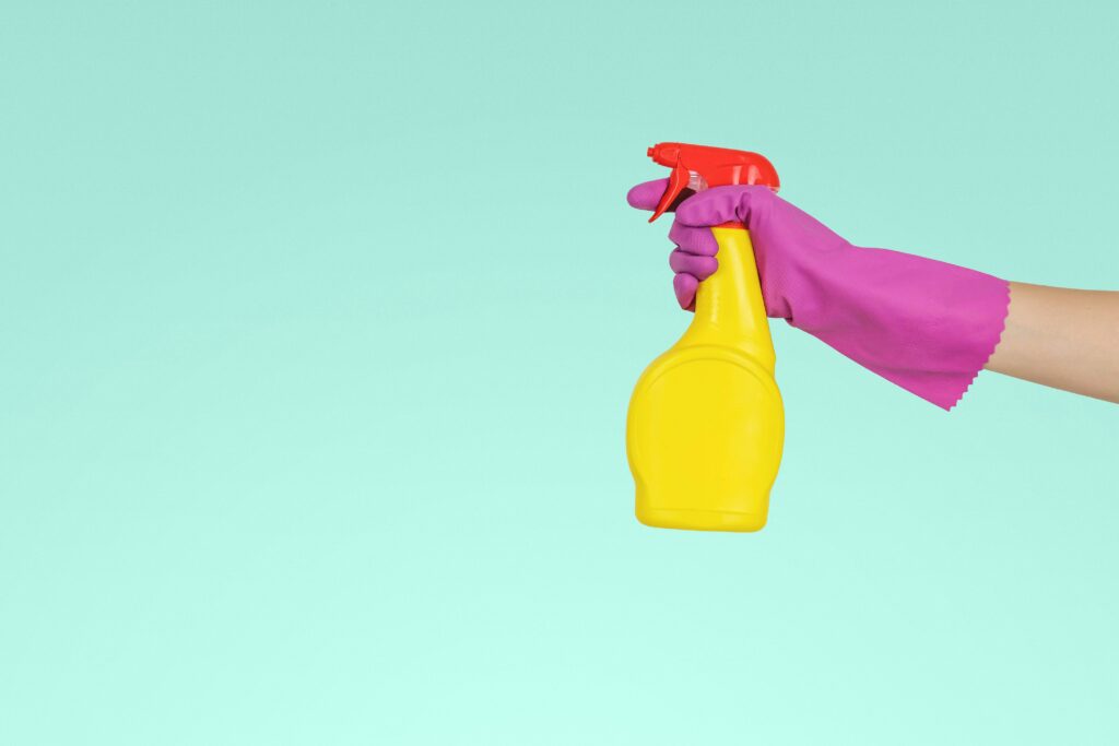 Hand in purple glove holding yellow spray bottle for cleaning against turquoise background.