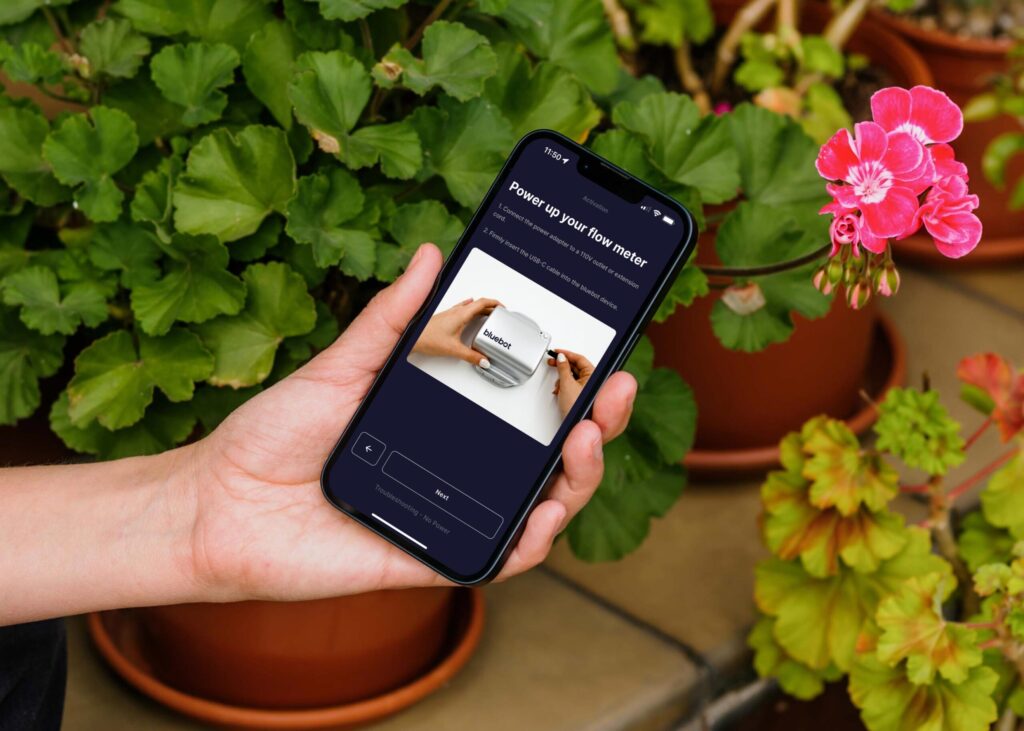 Person holding smartphone with flow meter instructions, surrounded by green and pink potted plants.