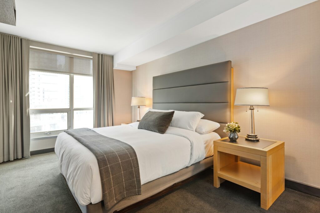 Modern hotel room with a comfortable bed, stylish headboard, bedside table with lamp and flowers, and large window.